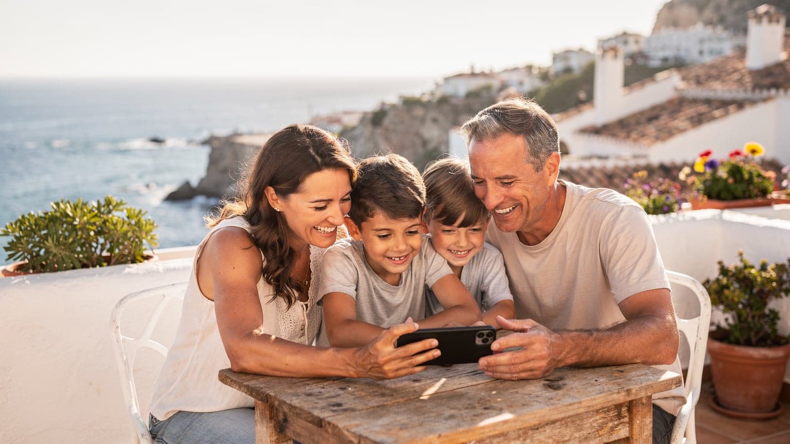 Una familia con dos niños, de unos 8 y 10 años, riendo mientras miran un teléfono móvil juntos en una terraza soleada con vistas al mar Mediterráneo en la Costa del Sol.