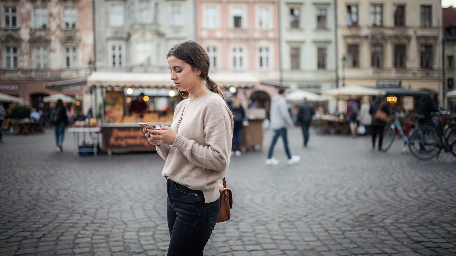Kobieta przeglądająca coś na telefonie na tle tętniącego życiem rynku w Krakowie, gdzie widać stragany z kwiatami i rękodziełem, a za nią w oddali charakterystyczne kamienice.
