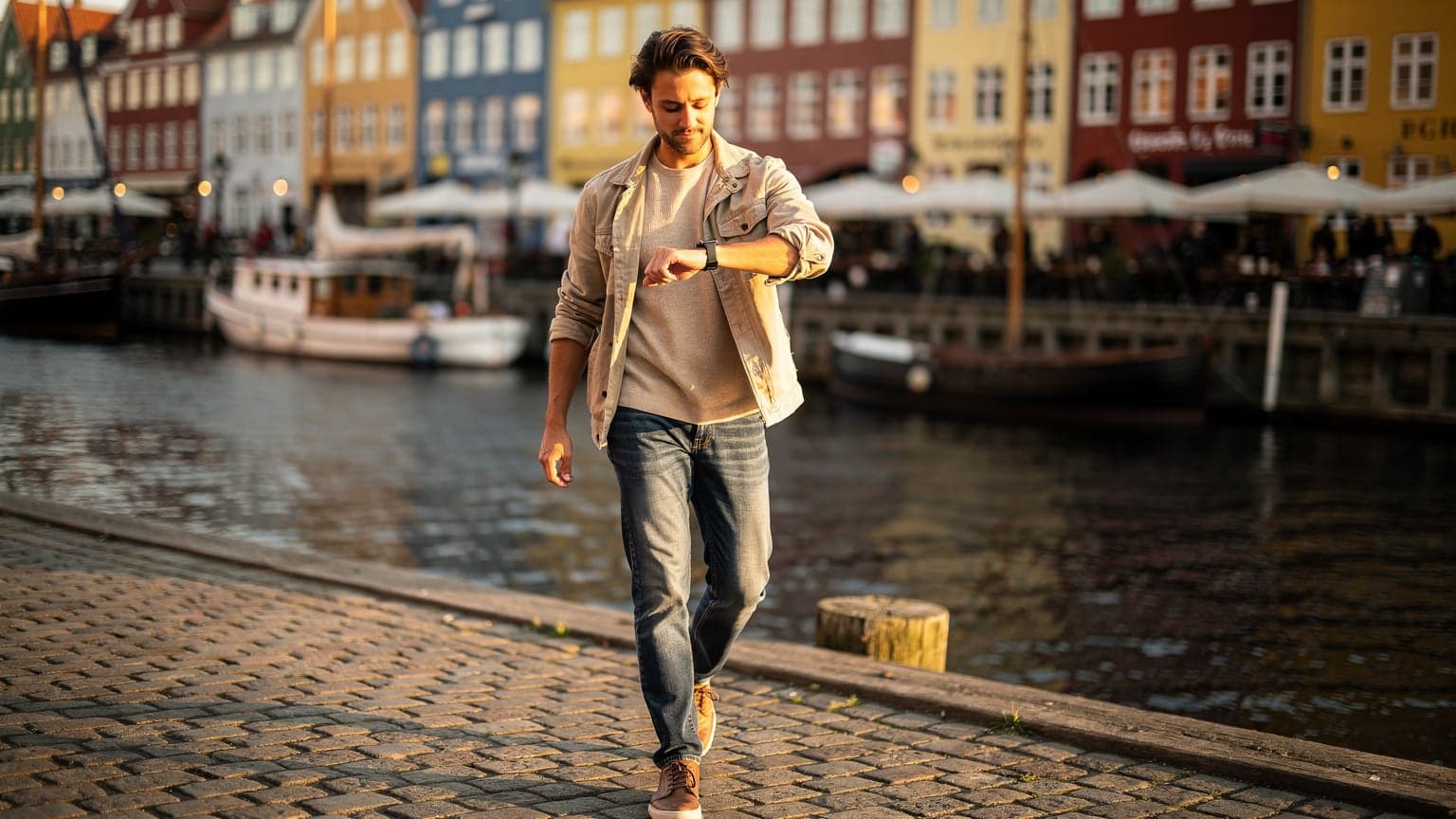 En person i Københavns indre by, iført let jakke, kigger på sit Apple Watch mens de går forbi Nyhavns farverige huse, solen skinner fra en klar himmel.
