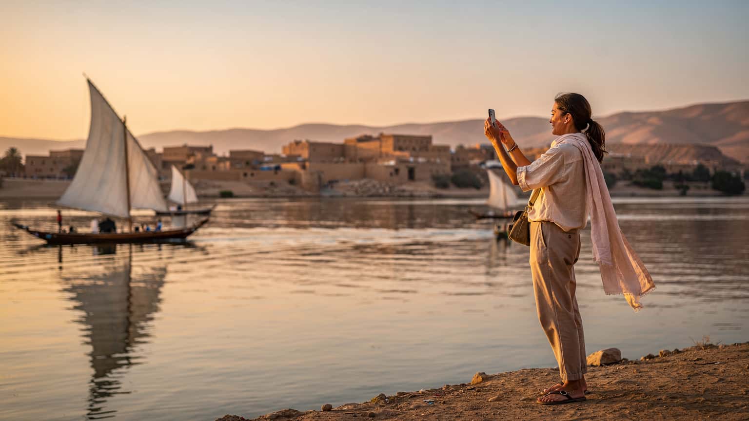 Une femme souriante, téléphone à la main, prend une photo du Nil au lever du soleil, avec des felouques traditionnelles en arrière-plan, près d'Assouan en Égypte.