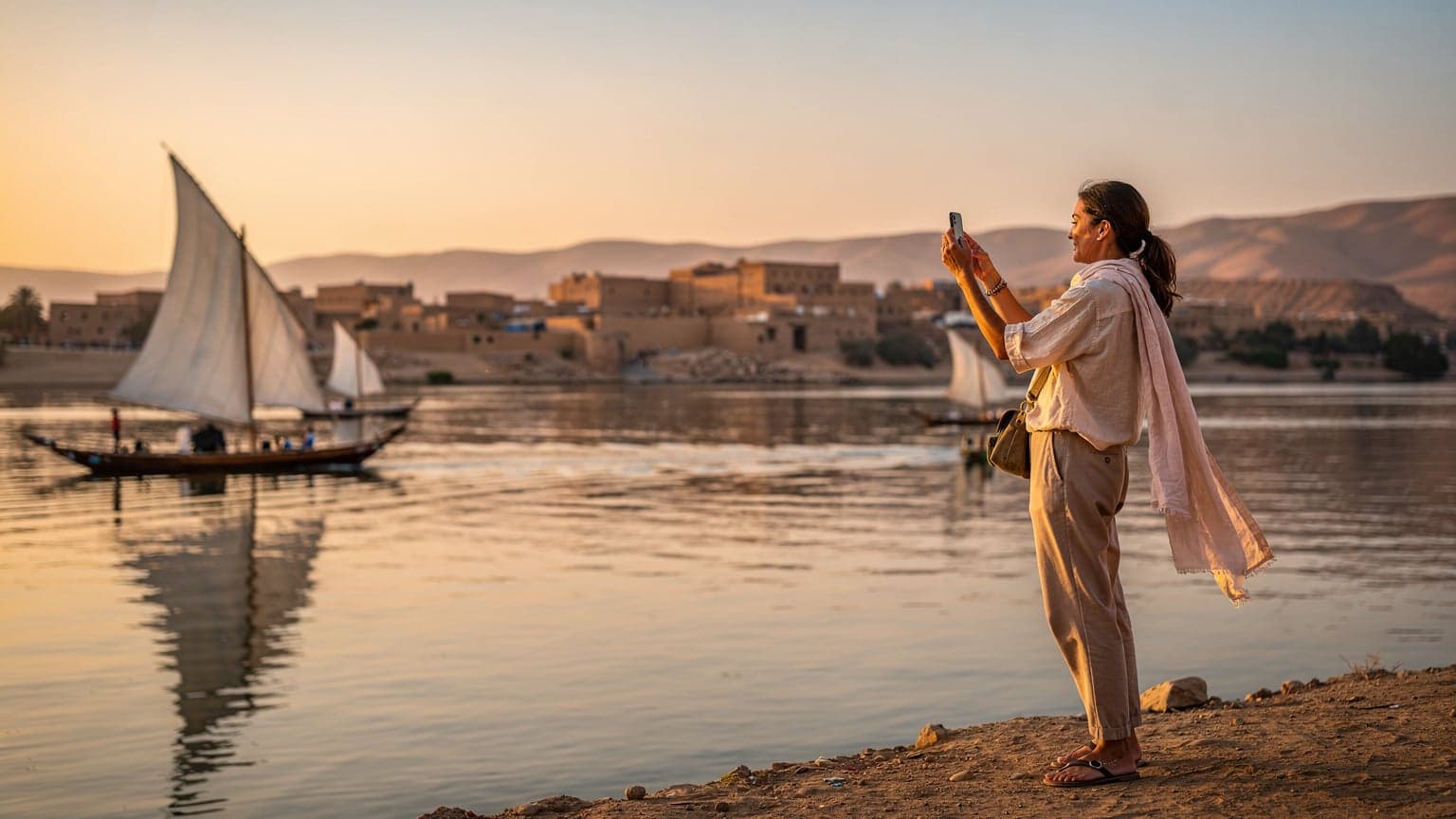 Une femme souriante, téléphone à la main, prend une photo du Nil au lever du soleil, avec des felouques traditionnelles en arrière-plan, près d'Assouan en Égypte.