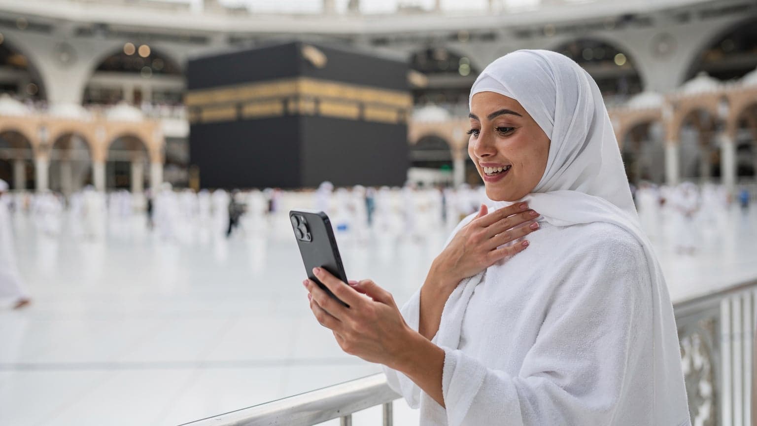 امرأة محجبة في الثلاثينات من عمرها تستخدم هاتفها الذكي داخل المسجد الحرام في مكة، تبتسم وتتحدث مع عائلتها عبر مكالمة فيديو، وتظهر الكعبة المشرفة في الخلفية بوضوح.