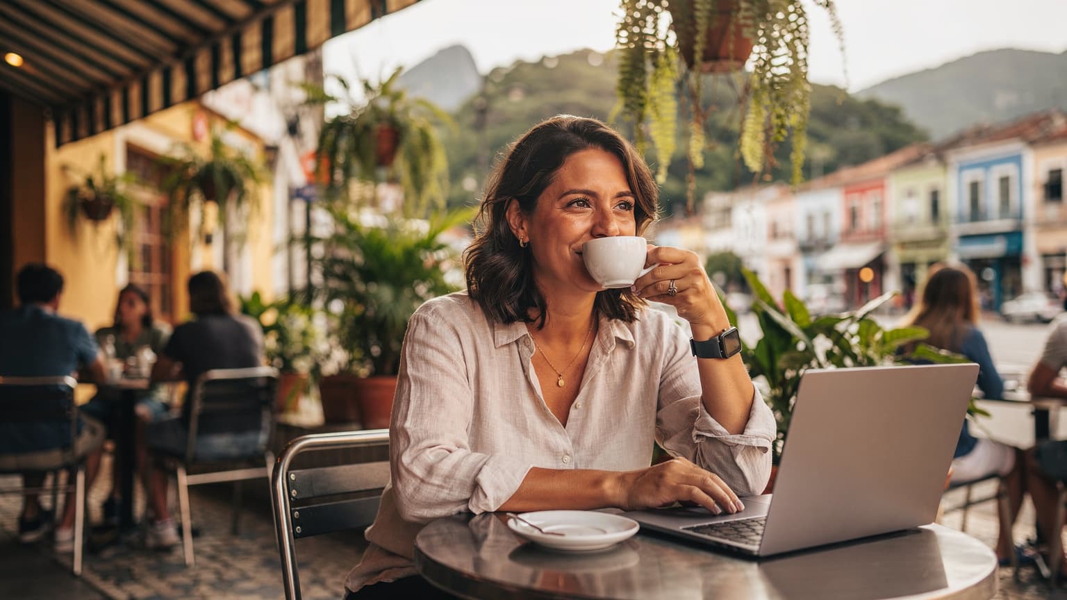 Una nomade digitale sorride mentre sorseggia un caffè caldo in un bar all'aperto di Rio de Janeiro, con una vista parziale della città in sottofondo.