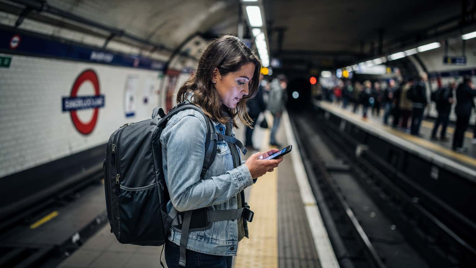 Een vrouw met een rugzak kijkt op haar telefoon op een drukke stationshal in Londen, met de herkenbare rode Backerloo Line borden op de achtergrond.