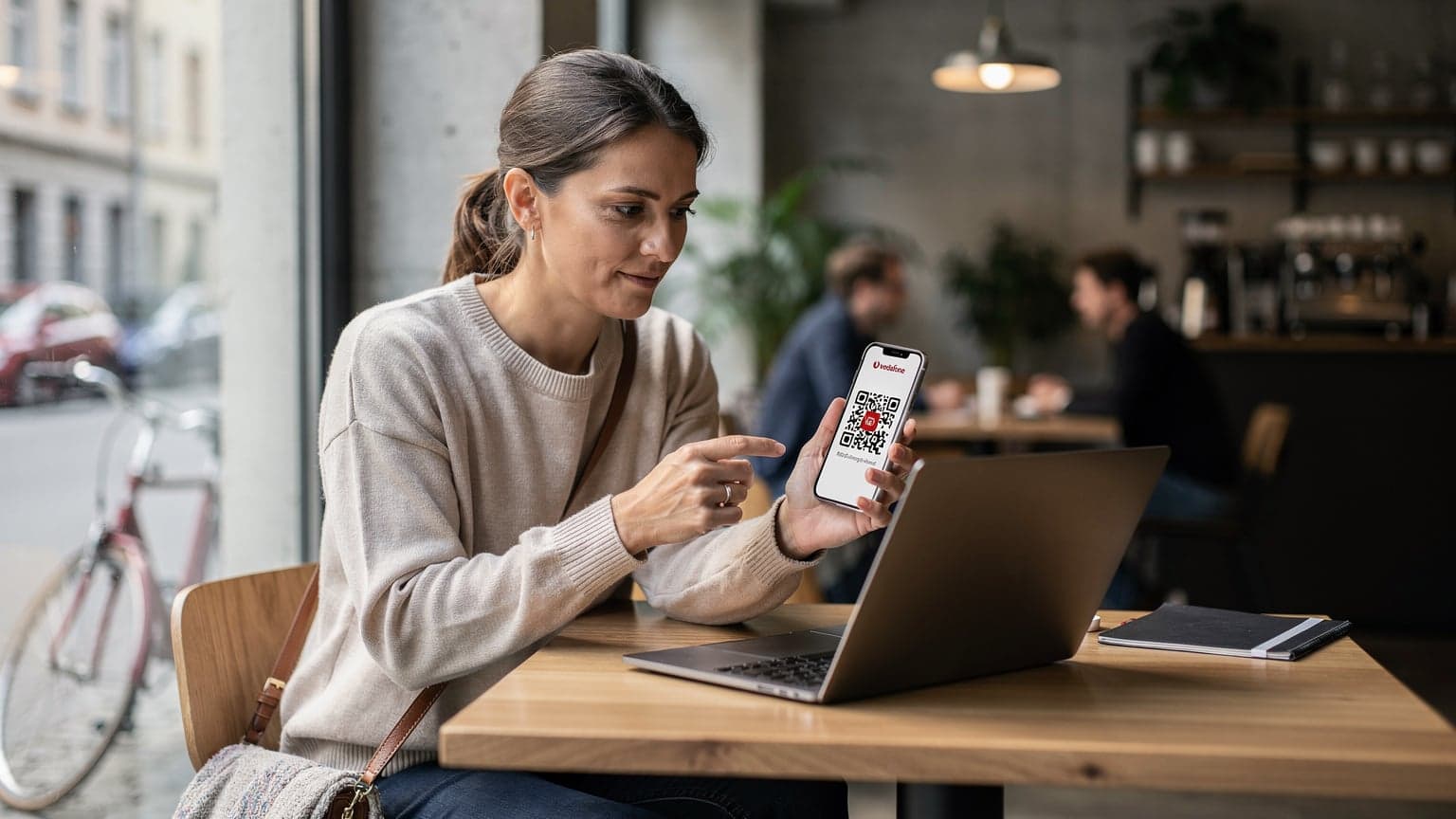 Eine Frau sitzt entspannt in einem Münchner Café mit Laptop und Smartphone, während sie eine Vodafone eSIM für ihre nächste Reise aktiviert.