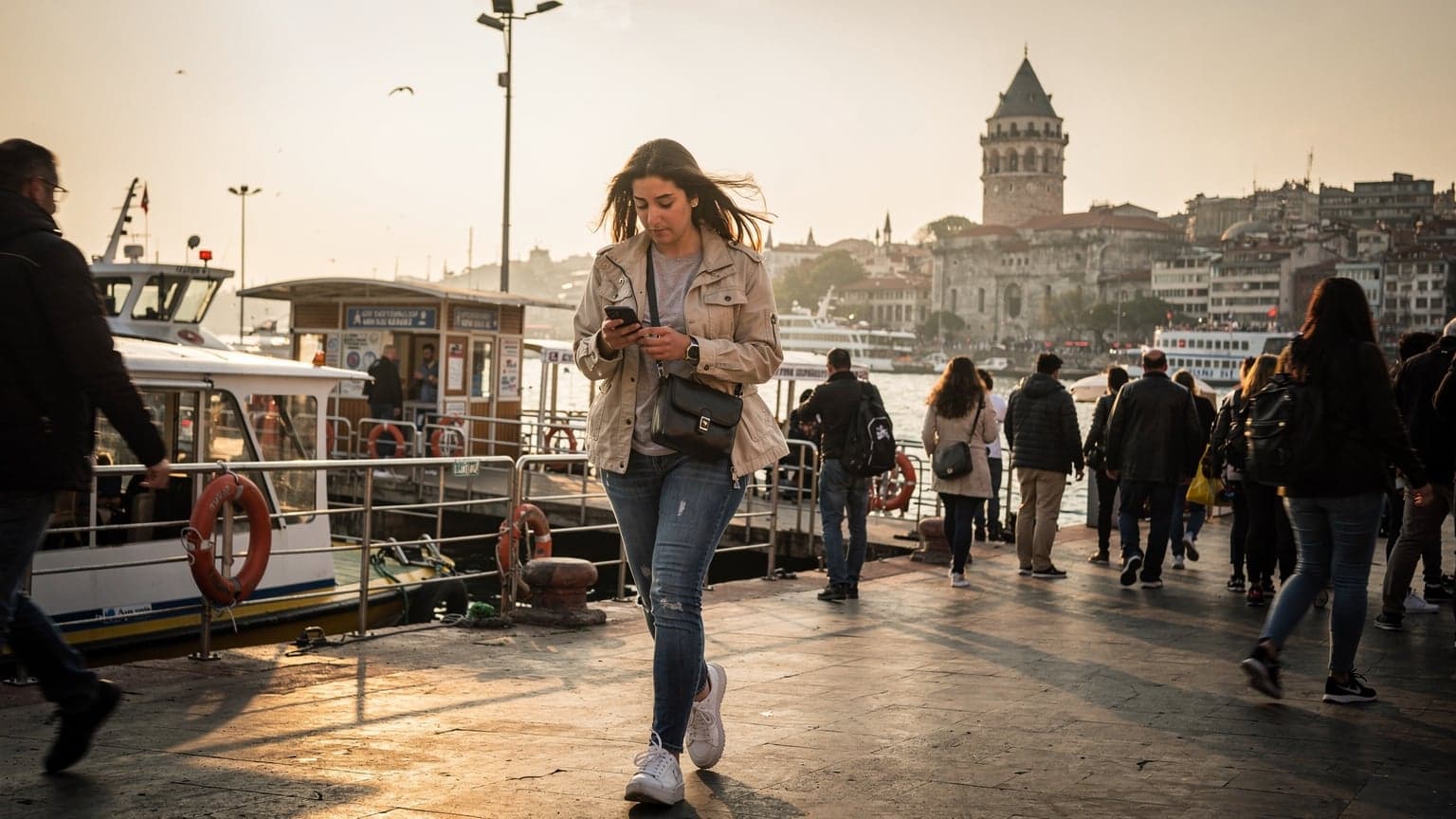 A woman confidently navigating the bustling Eminönü ferry terminal in Istanbul, holding her smartphone, with the iconic Galata Tower visible in the background under a warm afternoon sun.