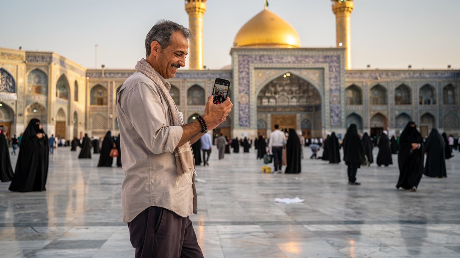 یک زائر میانسال فارسیزبان در حال قدم زدن در صحن حرم امام علی (ع) در نجف اشرف, با موبایل خود در حال برقراری تماس تصویری با خانواده است. او لبخندی بر لب دارد و پسزمینه, معماری اسلامی و خیل جمعیت را نشان میدهد.