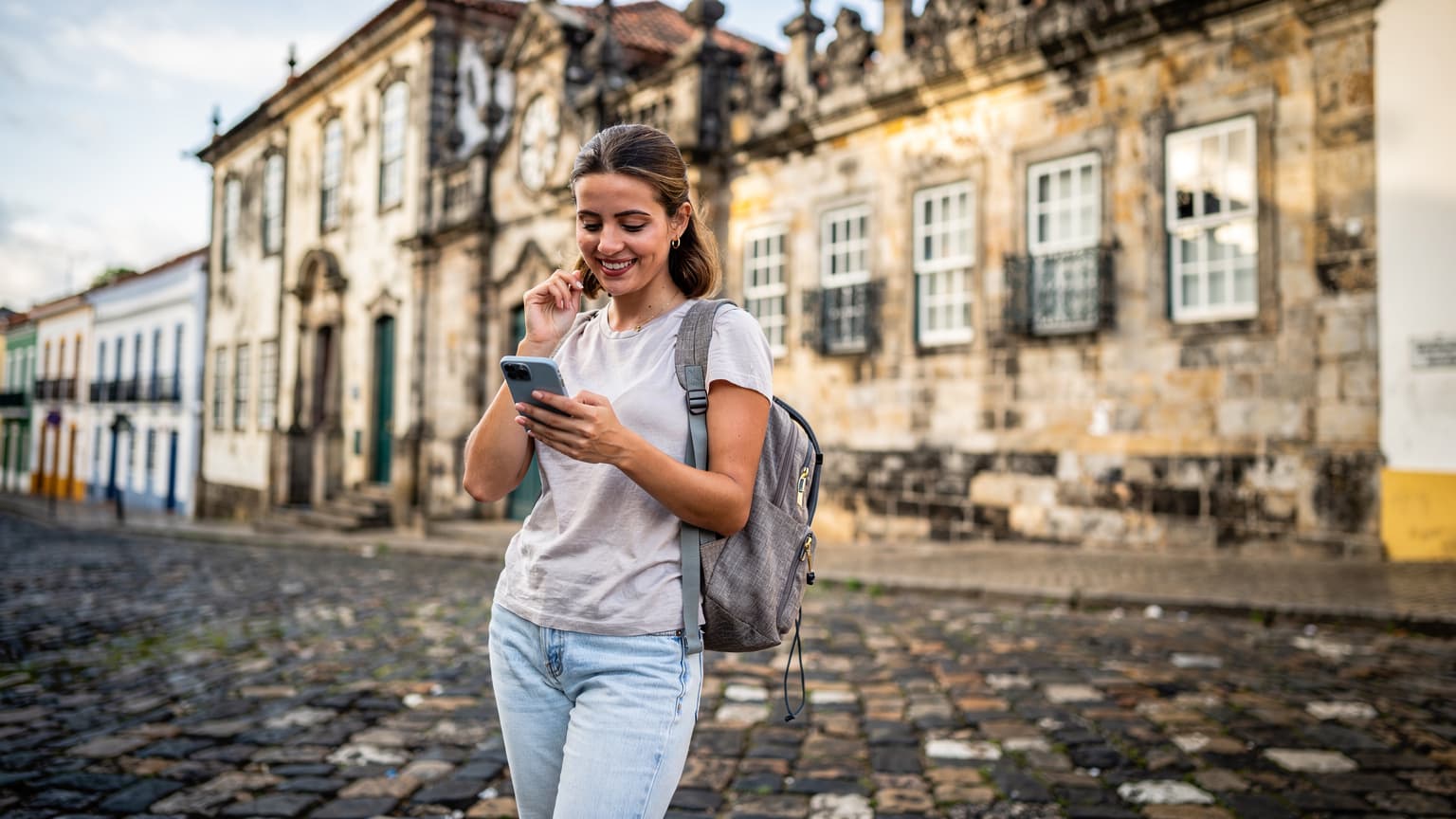 Mulher sorridente usa seu smartphone na frente do Mosteiro de São Bento em Salvador, Bahia, durante o dia, com luz solar suave iluminando a arquitetura colonial.