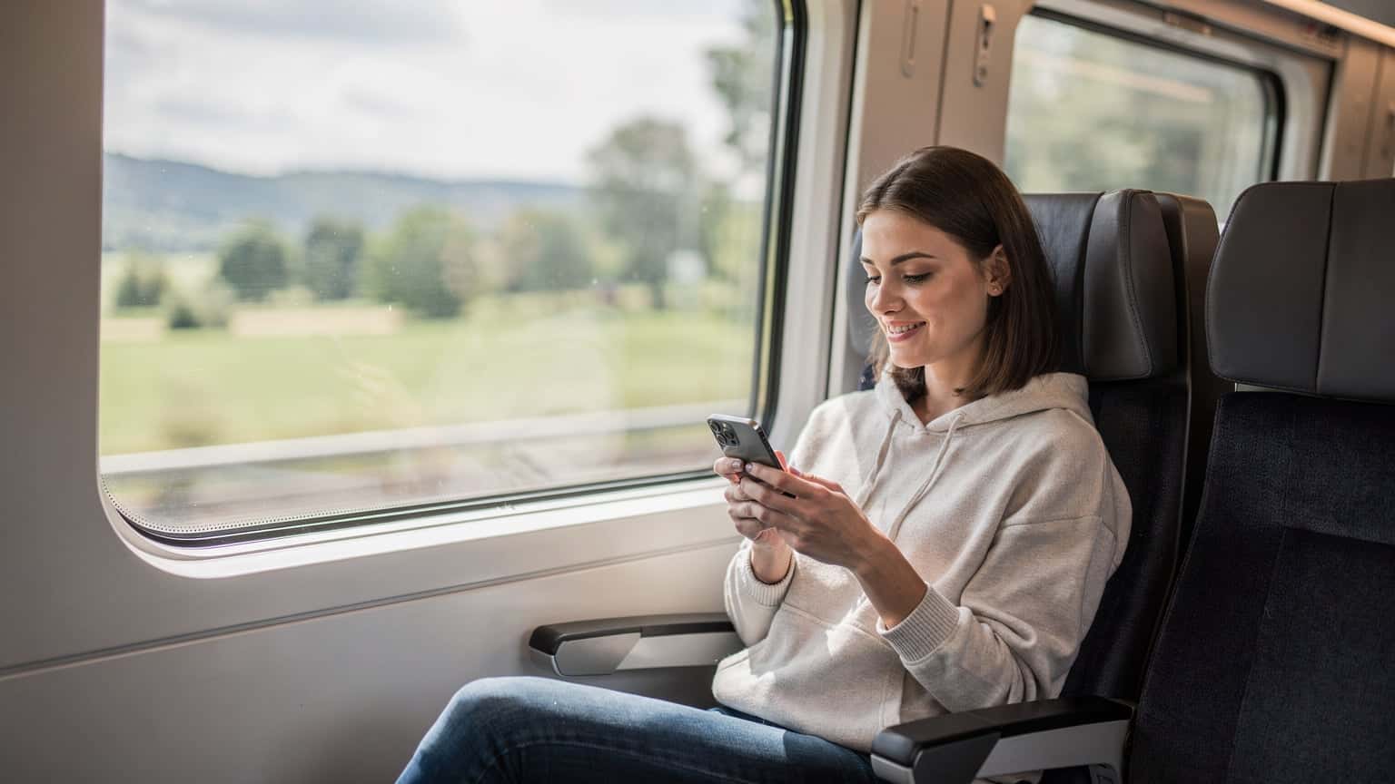 En ung kvinde sidder i et moderne europæisk tog med udsigt til forbipasserende landskab, hun kigger på sin telefon med et smil, mens hun planlægger sin rute.