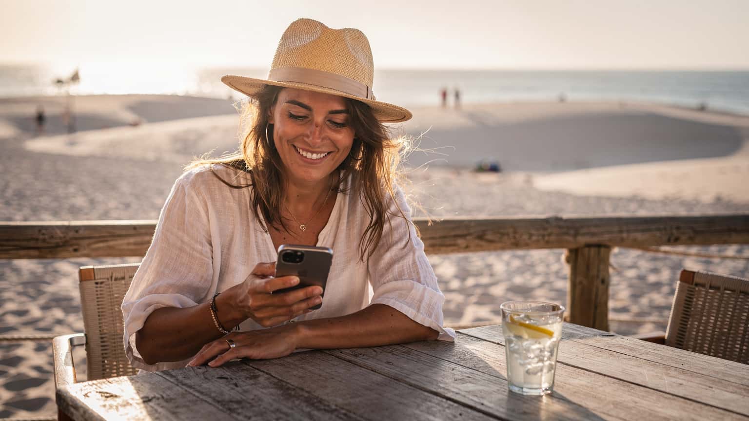 Una mujer sonriente, usando un sombrero de paja, consulta su smartphone en la terraza de un chiringuito en la playa de Bolonia, Cádiz, con las dunas al fondo y el mar Mediterráneo brillando bajo el sol de la tarde.