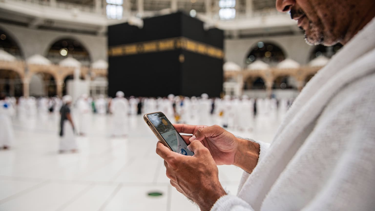 حاج مصري يرتدي الإحرام ويتصفح هاتفه الذكي داخل الحرم المكي الشريف، مع خلفية للكعبة المشرفة والناس يطوفون حولها.