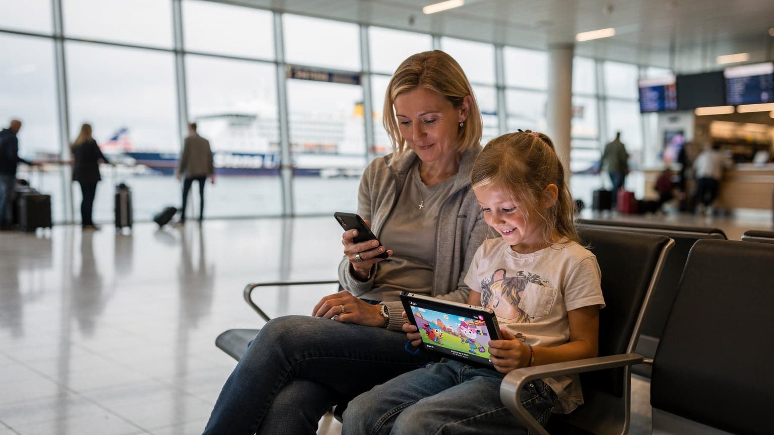 En mamma med en glad 8-årig dotter och en surfplatta på en färjeterminal i Göteborg, med en Cellesim-logotyp synlig i bakgrunden.