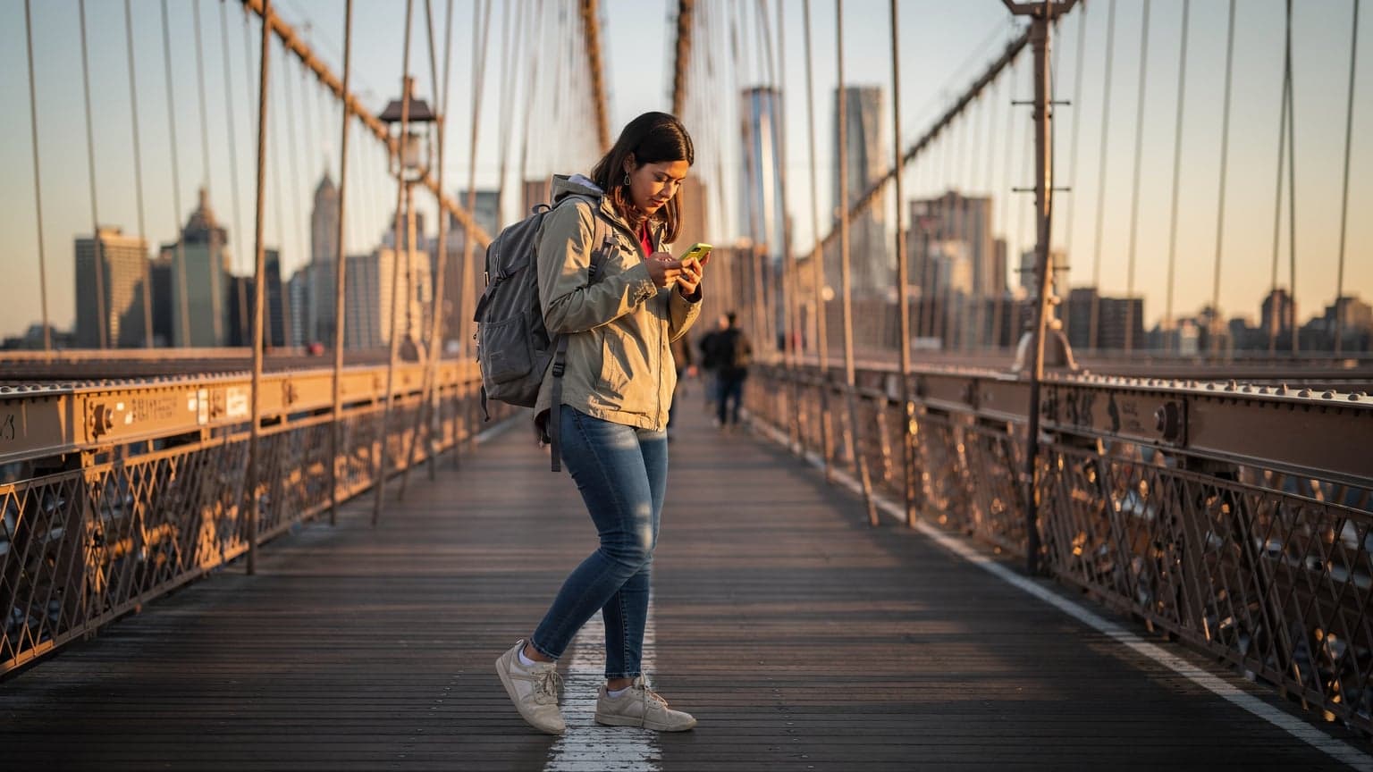 Mladá žena s batohem prochází po Brooklynském mostě v New Yorku a kontroluje telefon s výhledem na panorama Manhattanu za úsvitu.