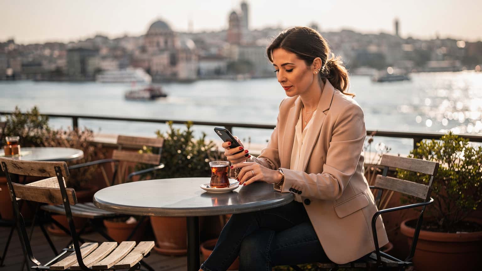 O femeie zâmbitoare navighează pe telefonul ei, cu o privire relaxată, în timp ce savurează un ceai turcesc pe o terasă însorită cu vedere la Bosfor, în cartierul Karaköy din Istanbul.