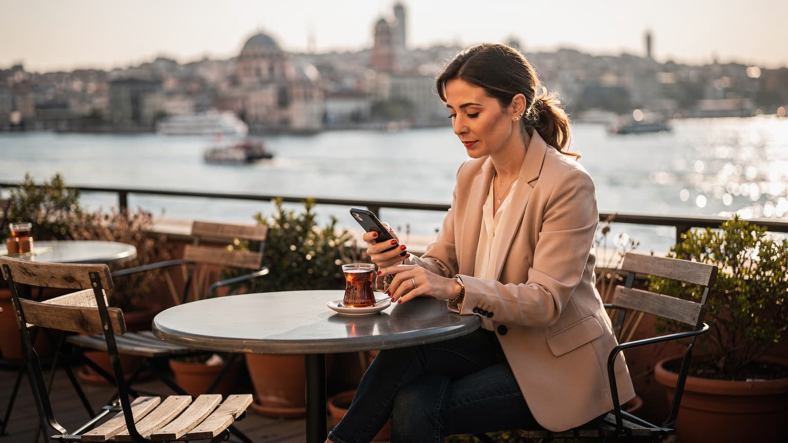 O femeie zâmbitoare navighează pe telefonul ei, cu o privire relaxată, în timp ce savurează un ceai turcesc pe o terasă însorită cu vedere la Bosfor, în cartierul Karaköy din Istanbul.