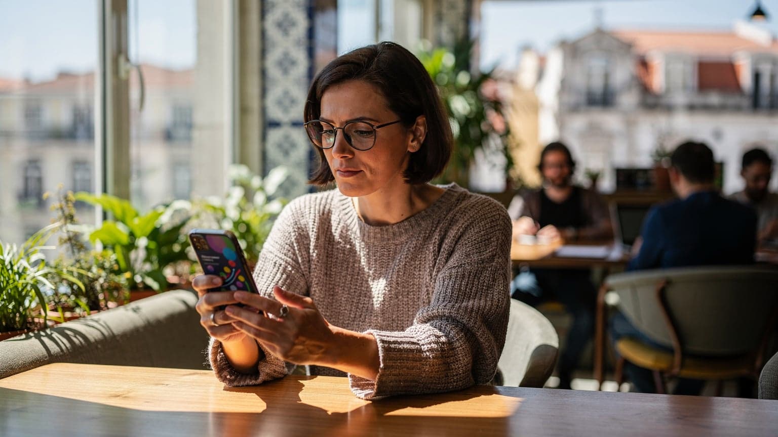 A digital nomad sits at a wooden table in a busy co-working space in Lisbon, Portugal, looking intently at her phone with an open laptop beside her, a half-empty coffee cup in hand.