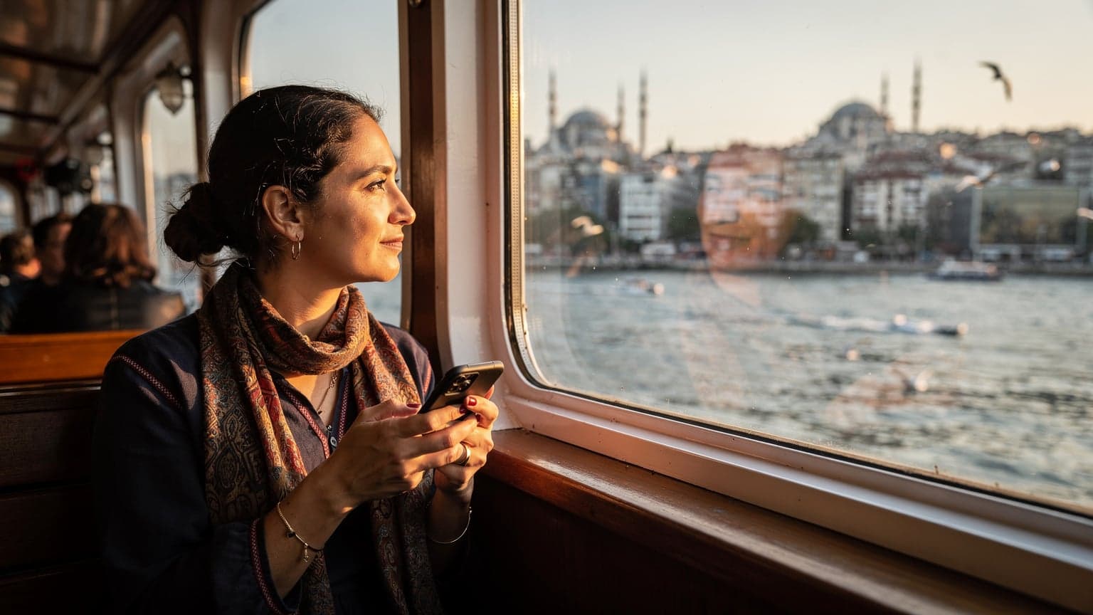 İstanbul Boğazı'nda bir vapurda oturan, elinde telefonunu tutan bir kadın ufuk çizgisine bakıyor, şehir silüeti ve tarihi yapılar arka planda.