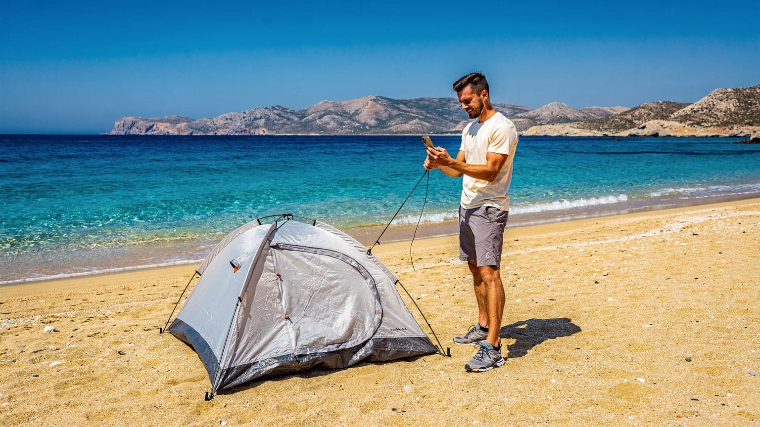 Νεαρός άνδρας στήνει σκηνή σε μια απομονωμένη, αμμώδη παραλία κάτω από τον λαμπερό ελληνικό ήλιο, με το κινητό του τηλέφωνο να δείχνει σήμα.