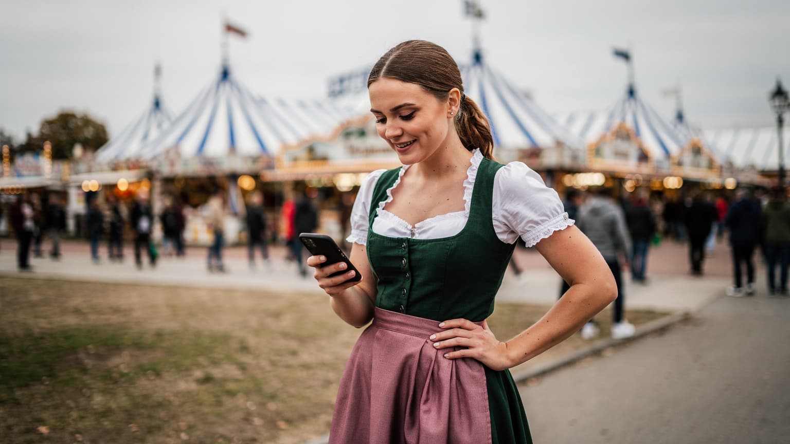 A woman in traditional Bavarian attire smiles, holding a smartphone showing a map, with the bustling beer tents of Oktoberfest in the background under a soft, overcast autumn sky.