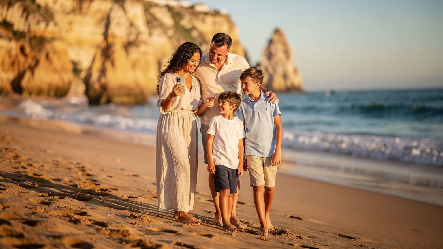 O familie zâmbitoare, părinți și doi copii cu vârste între 8 și 12 ani, se bucură de o plimbare pe malul oceanului la Praia da Rocha în Algarve, Portugalia, în timp ce consultă o hartă digitală pe un smartphone.
