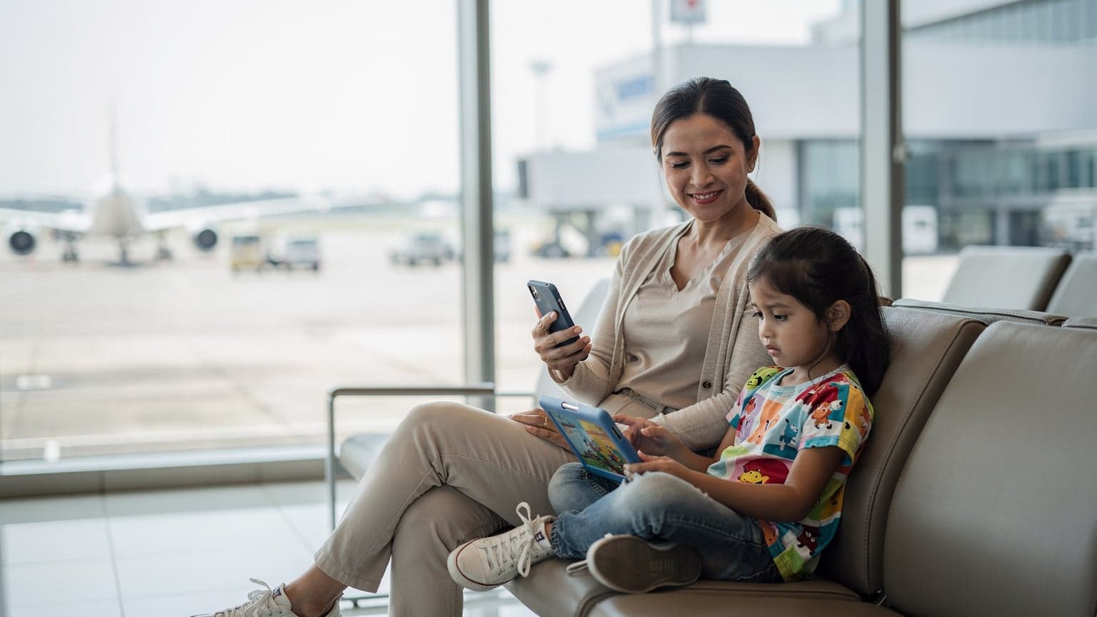 Seorang ibu tersenyum lega sambil memegang iPhone di tangan kirinya dan menggandeng tangan anak perempuannya yang sedang asyik menonton kartun di iPad di sebuah lounge keberangkatan Bandara Internasional Soekarno-Hatta yang modern dan terang.