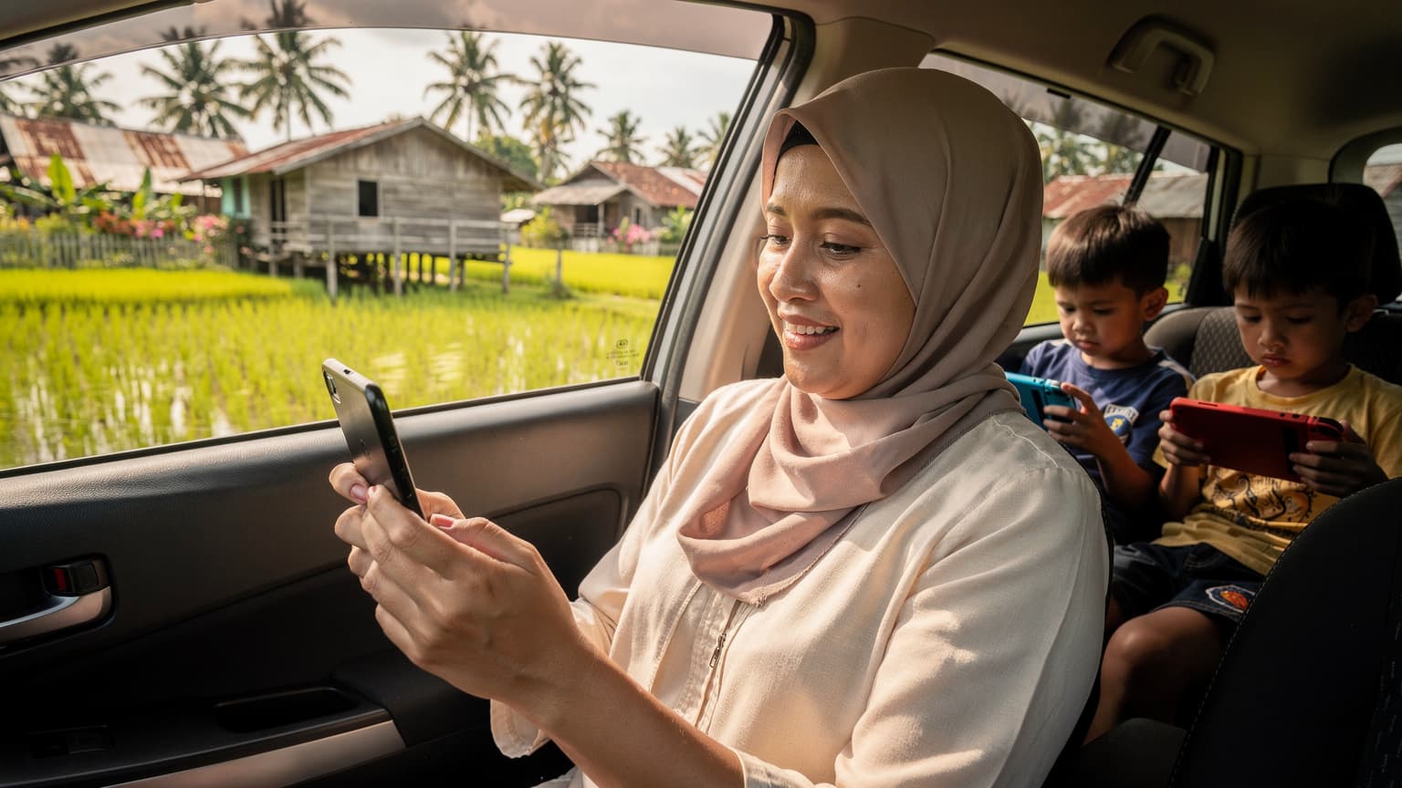 Seorang ibu tersenyum lega sambil melihat telefon bimbitnya yang menunjukkan isyarat rangkaian penuh, dikelilingi oleh anak-anaknya yang asyik dengan peranti masing-masing di dalam kereta yang bergerak pulang ke kampung, melalui landskap hijau luar bandar Malaysia.