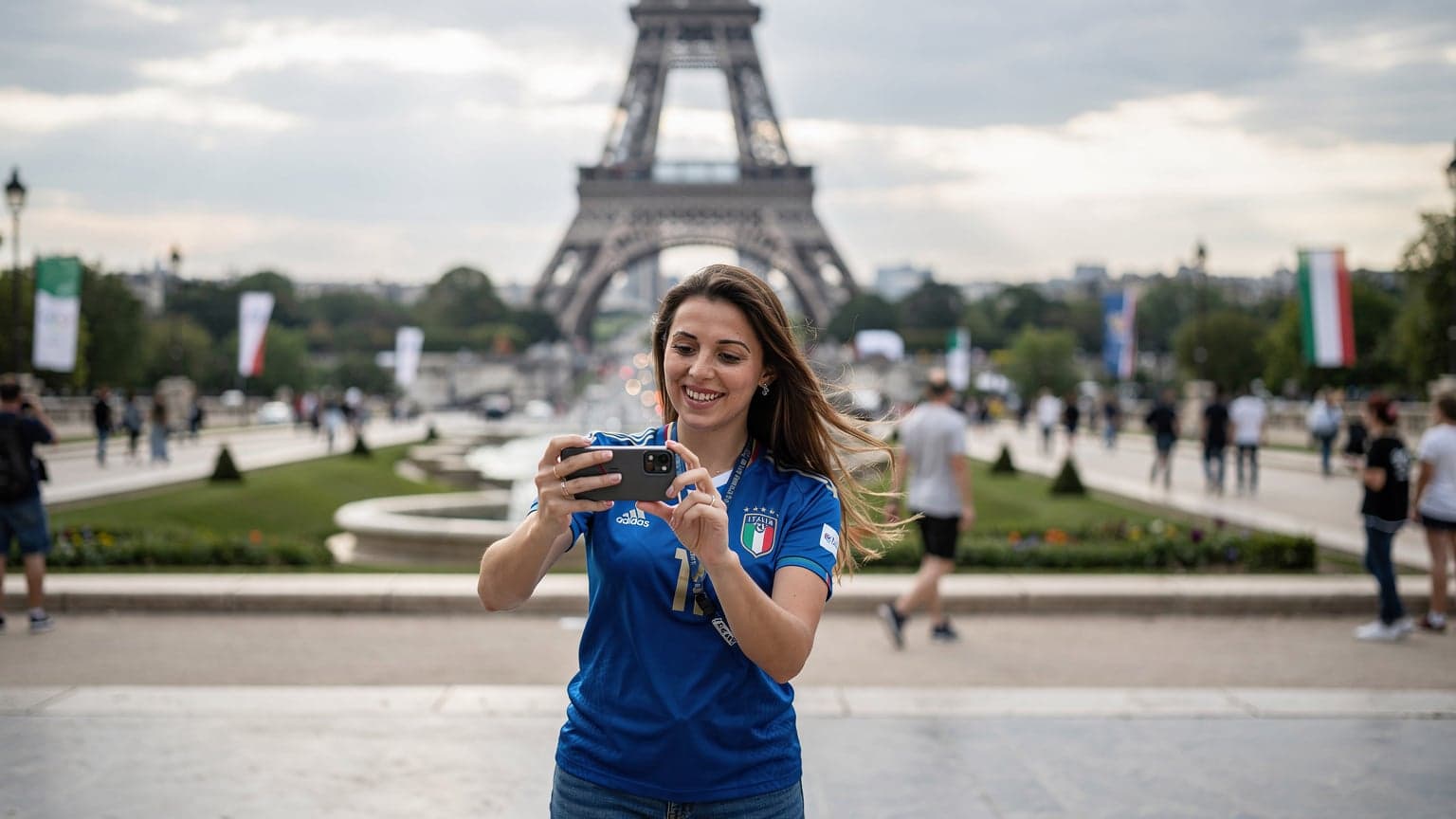 Una tifosa italiana sorridente con una maglia azzurra scatta una foto del Trocadéro con la Torre Eiffel sullo sfondo, usando il suo smartphone con un'eSIM attiva.