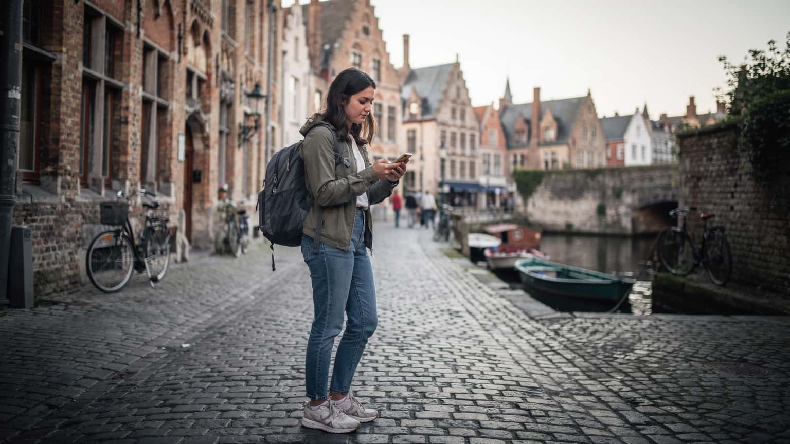 Eine junge Frau mit Rucksack und Smartphone navigiert durch die charmanten Gassen von Brügge, während sie auf ihr Handy schaut und die mittelalterliche Architektur bewundert.
