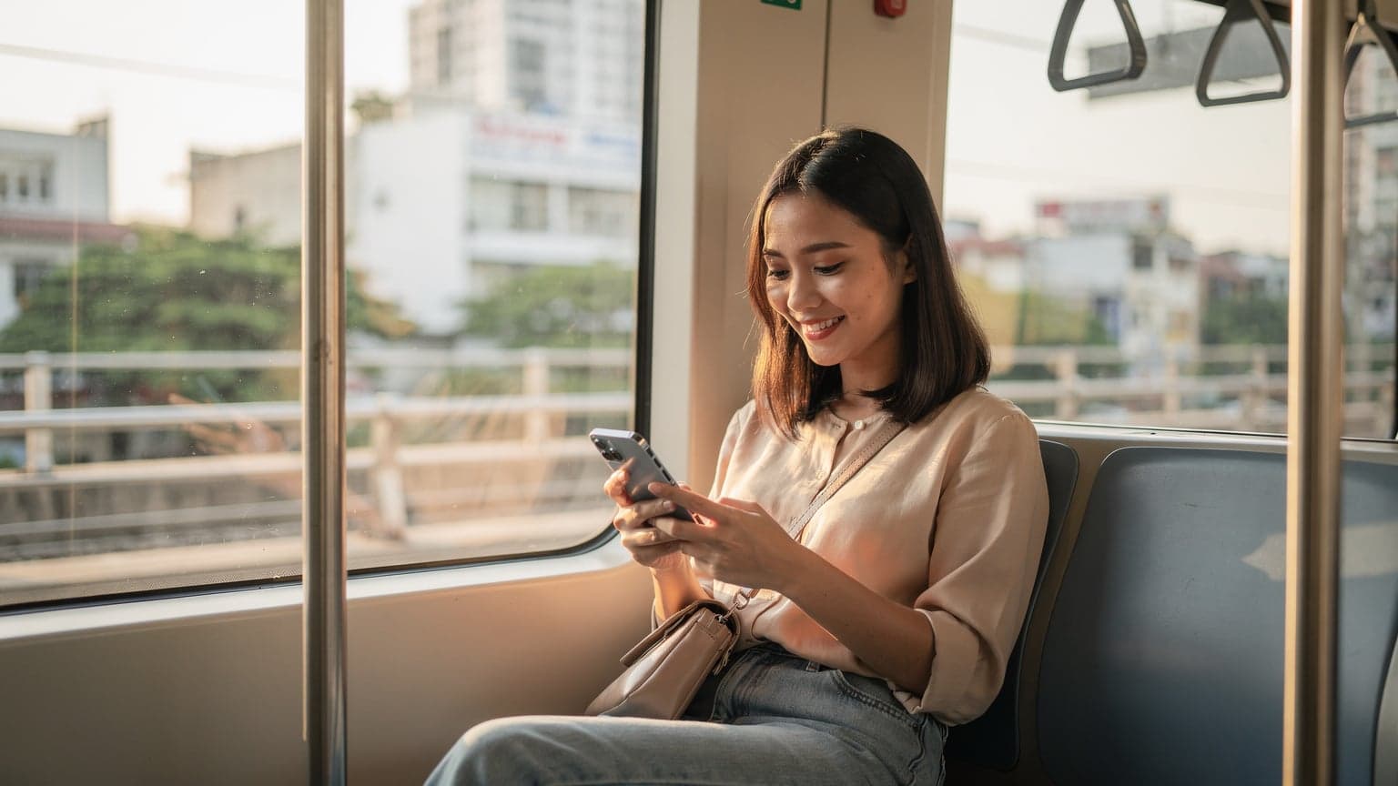 Một phụ nữ Việt Nam trẻ tuổi đang mỉm cười, cầm trên tay chiếc iPhone đã bật màn hình trong khi ngồi trên chuyến tàu điện ngầm tuyến số 1 tại Thành phố Hồ Chí Minh, với ánh nắng ban mai chiếu qua cửa sổ.
