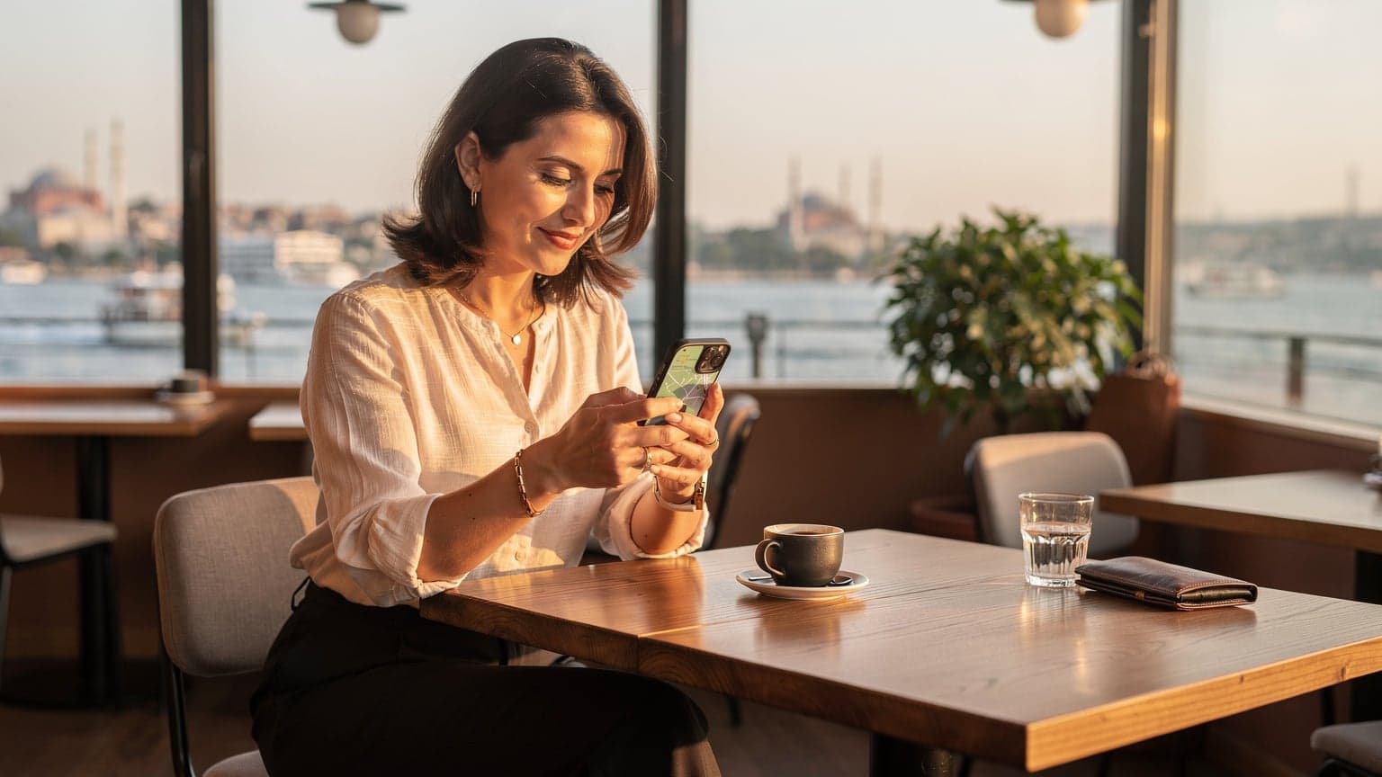 O femeie zâmbitoare, cu un iPhone 15 în mână, verificând harta într-o cafenea modernă cu vedere spre Bosfor în Istanbul, pe fundal fiind vizibile minaretele Hagiei Sofia.