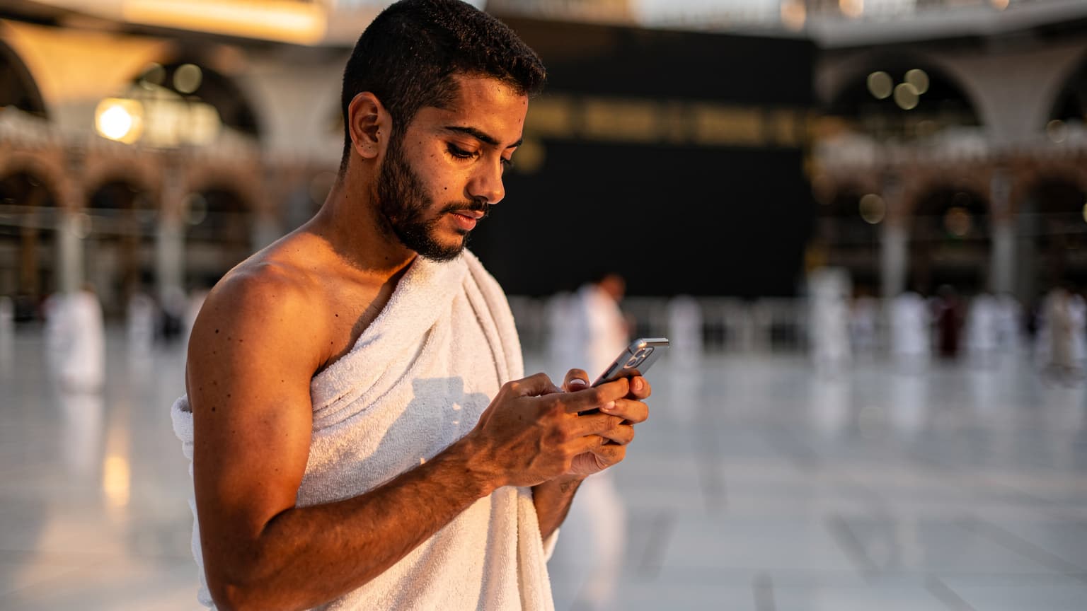 معتمر شاب من مصر يتصفح هاتفه الذكي داخل الحرم المكي الشريف، يظهر الكعبة المشرفة في الخلفية، مع إضاءة دافئة وقت المغرب.