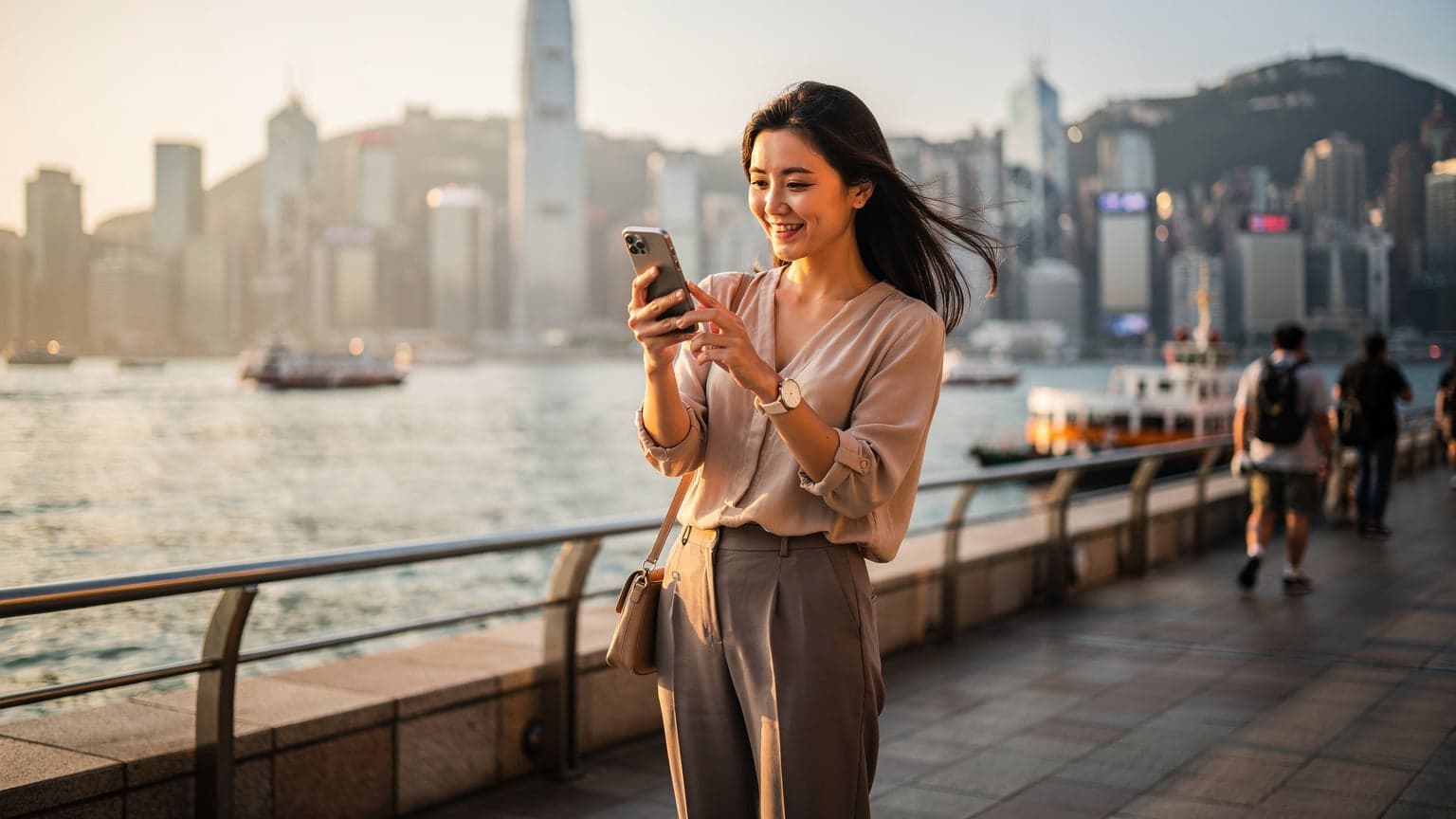 一名年轻女士手持iPhone,站在香港维多利亚港湾繁忙的尖沙咀海滨,背景是香港岛的摩天大楼和天星小轮,她正在屏幕上查看信息。