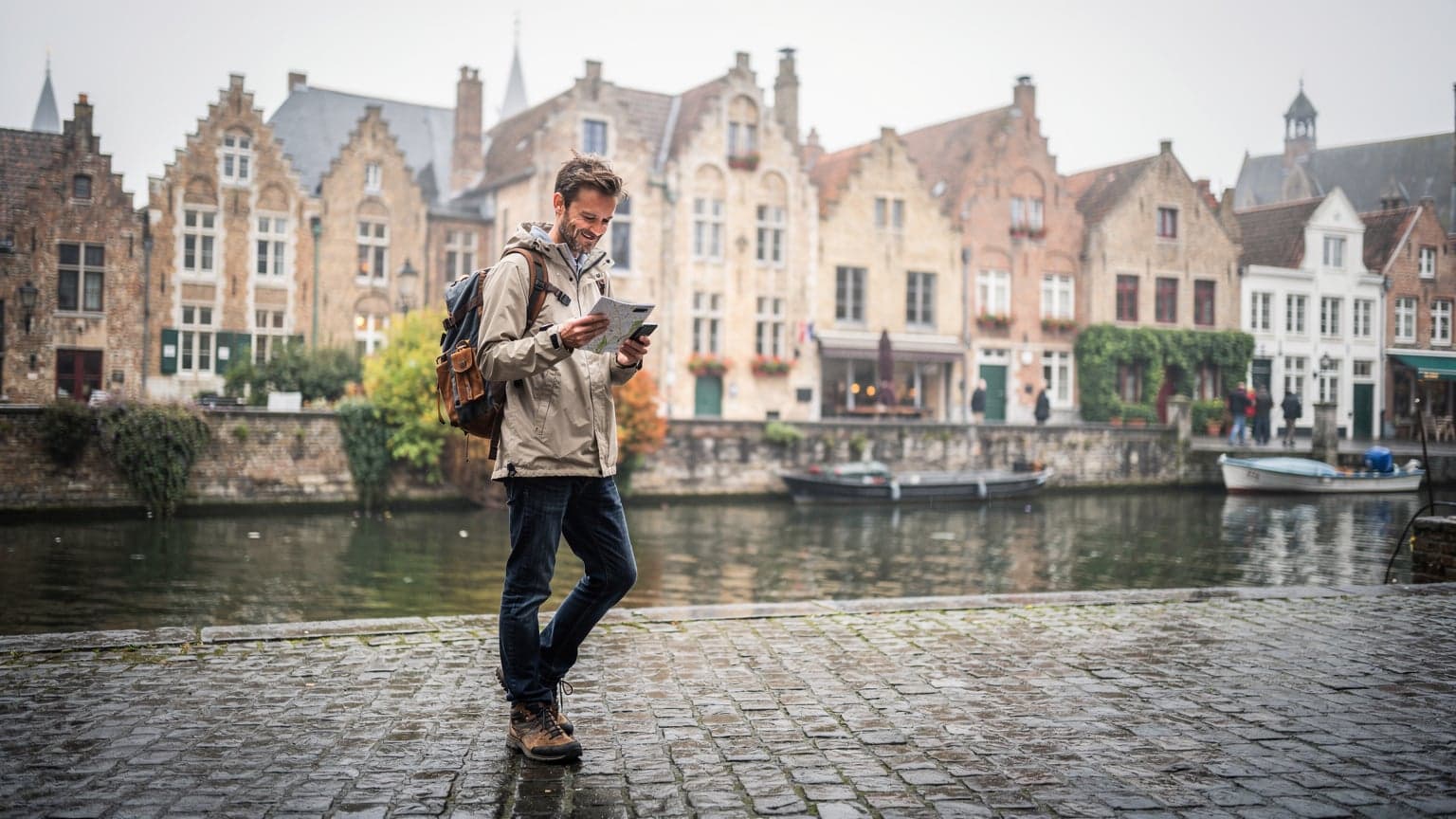 Un voyageur souriant, sac à dos, debout sur une petite place pavée de Bruges, avec des maisons à pignons typiques en arrière-plan et un canal reflétant le ciel nuageux.