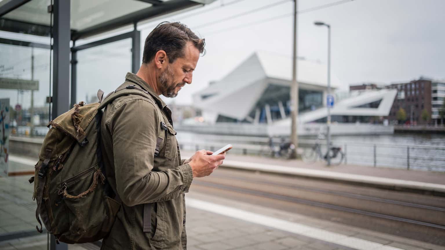 Een persoon met een rugzak kijkt op hun smartphone naar een kaart terwijl ze op een tramhalte in Amsterdam-Noord staan, met moderne architectuur op de achtergrond.