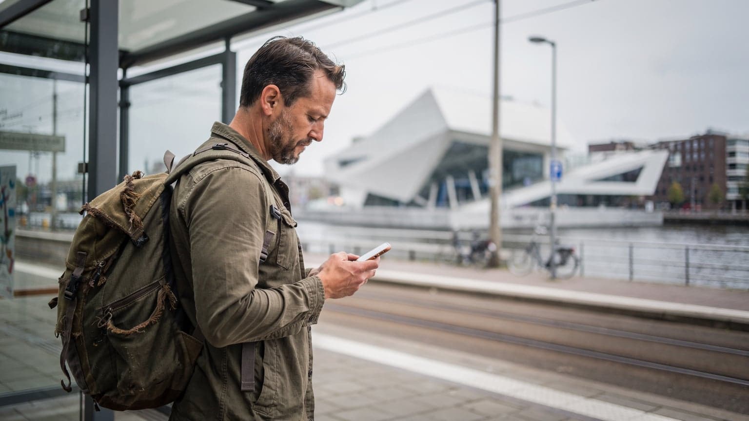 Een persoon met een rugzak kijkt op hun smartphone naar een kaart terwijl ze op een tramhalte in Amsterdam-Noord staan, met moderne architectuur op de achtergrond.