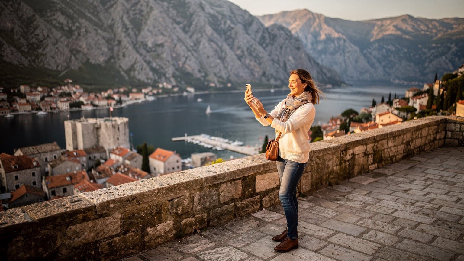 Una donna sorride mentre utilizza il suo smartphone su una terrazza panoramica che si affaccia sulla Baia di Kotor in Montenegro, con le montagne e le acque blu sullo sfondo al tramonto.