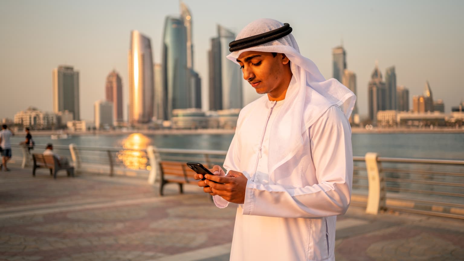 شاب يرتدي غترة ويستخدم هاتفه الذكي على كورنيش المنامة المطل على الأفق، مع أبراج خليج البحرين وناطحات السحاب في الخلفية، تحت سماء صافية عند الغروب.