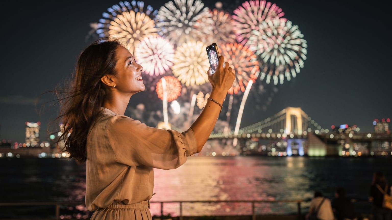 Seorang wanita muda memegang ponsel di tangannya, merekam festival kembang api Hanabi yang spektakuler di atas Teluk Tokyo pada malam hari, dengan siluet jembatan dan gedung-gedung tinggi terlihat di kejauhan.