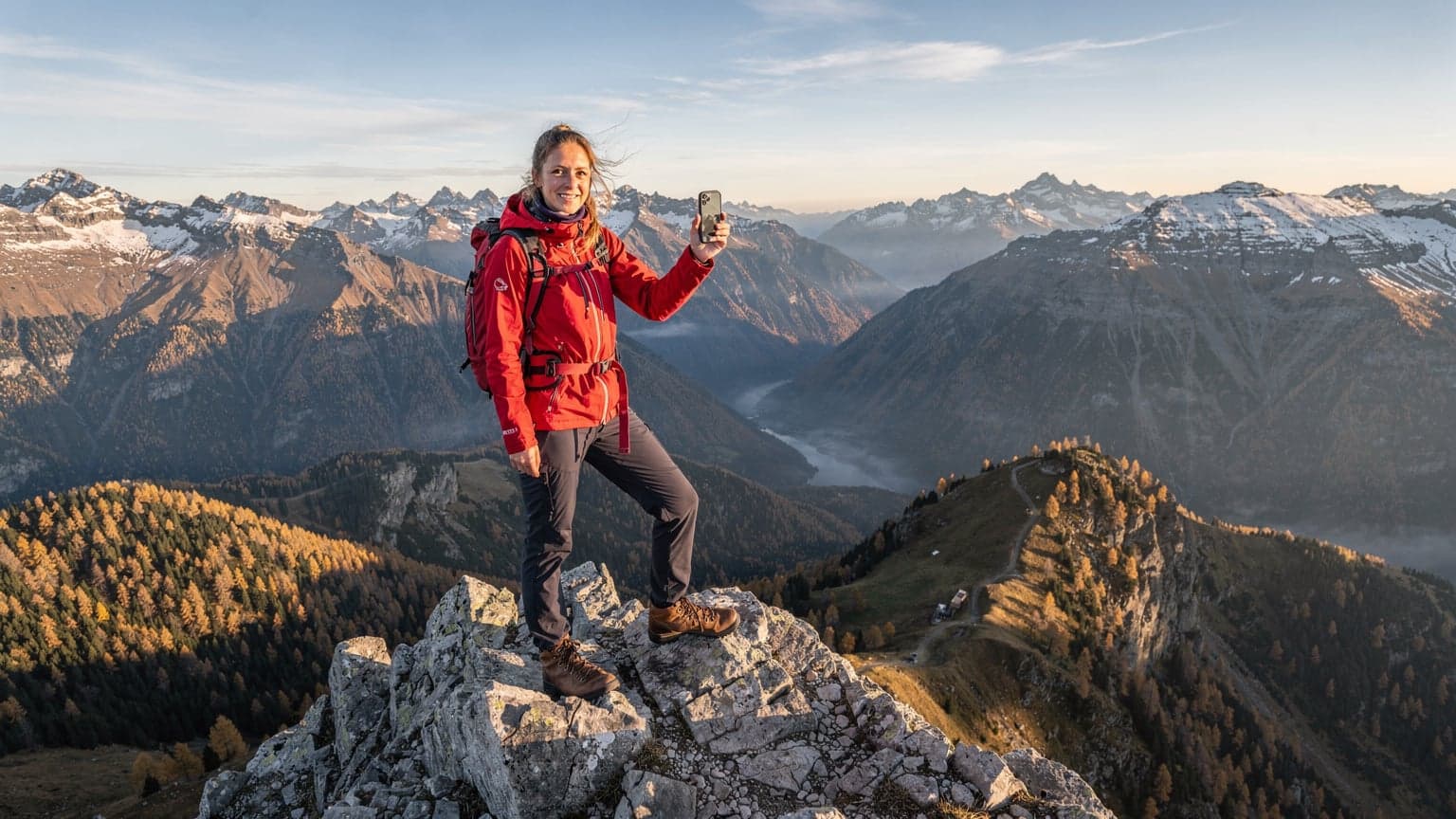 Seorang pendaki wanita berjaket hiking merah sedang berpose di puncak gunung di Pegunungan Alpen Eropa saat musim gugur, dengan smartphone di tangan menunjukkan sinyal penuh, latar belakang pemandangan lembah dan gunung bersalju yang indah.