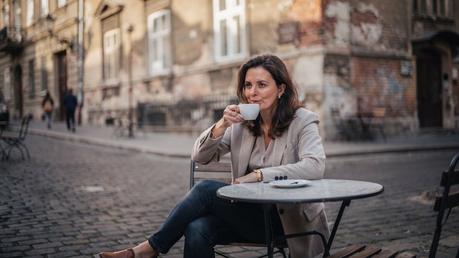 A woman sips coffee at an outdoor cafe table in Budapest's Jewish Quarter, a historic synagogue visible in the background under soft morning light.