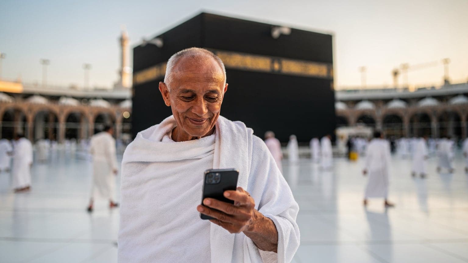 Seorang jemaah haji yang lebih tua sedang melihat telefonnya dengan senyuman, di latar belakang Kaabah di Makkah pada waktu subuh, menunjukkan sambungan yang lancar.