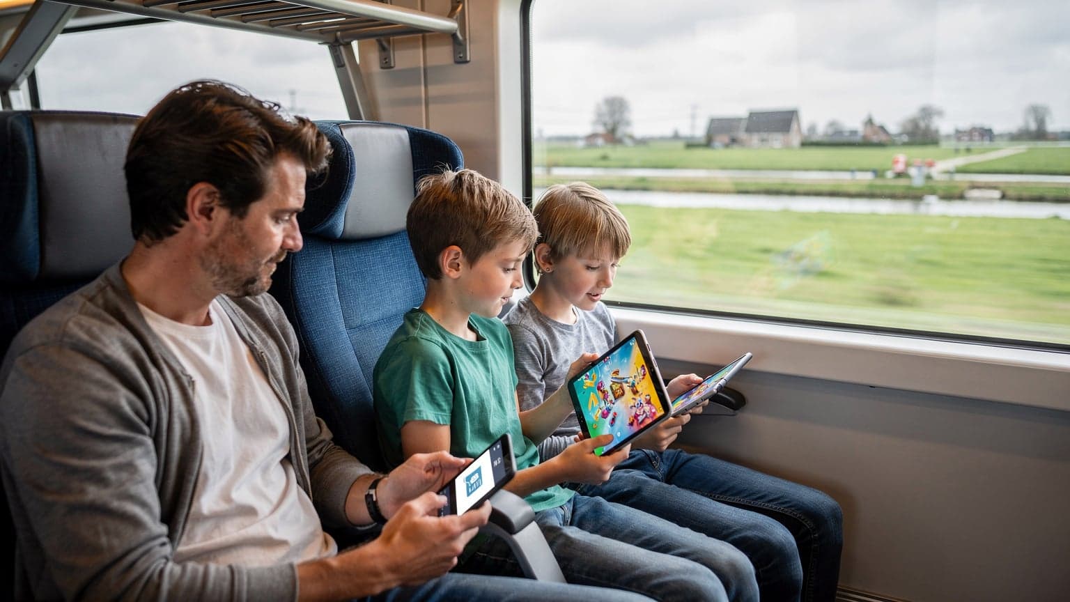 Een familie met twee kinderen, elk met een tablet of telefoon, zit ontspannen in een treincoupé in Nederland terwijl ze naar een landschap buiten kijken, waarbij de ouders glimlachen en de connectiviteit van hun apparaten controleren.