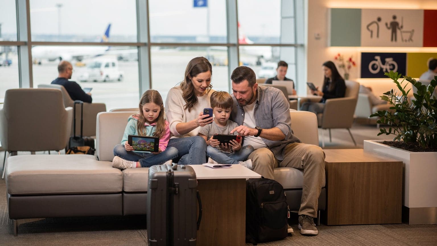 A family, two parents and two children aged around 8 and 10, are sitting in a bustling international airport lounge, engaged with their devices. The mother is showing something on her phone to her daughter, who has an iPad open on her lap playing a cartoon, while the father is helping his son with a Nintendo Switch. Luggage is neatly stacked nearby.