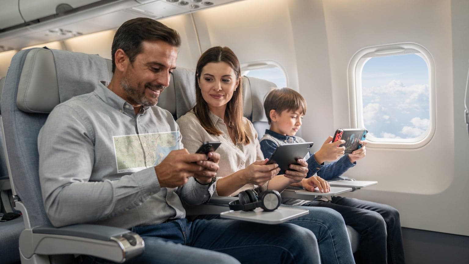 Uma família, pai, mãe e duas crianças, sentados confortavelmente num avião moderno, cada um com um dispositivo diferente, utilizando conectividade eSIM para entretenimento durante o voo.