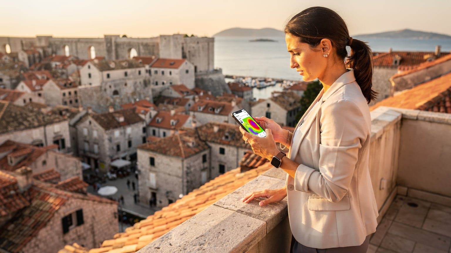 Una donna ingegnere delle telecomunicazioni con il suo smartphone in mano, sta visualizzando una mappa di copertura 5G mentre si trova su una terrazza panoramica che affaccia sulla città vecchia di Dubrovnik, Croazia, durante l'ora d'oro.