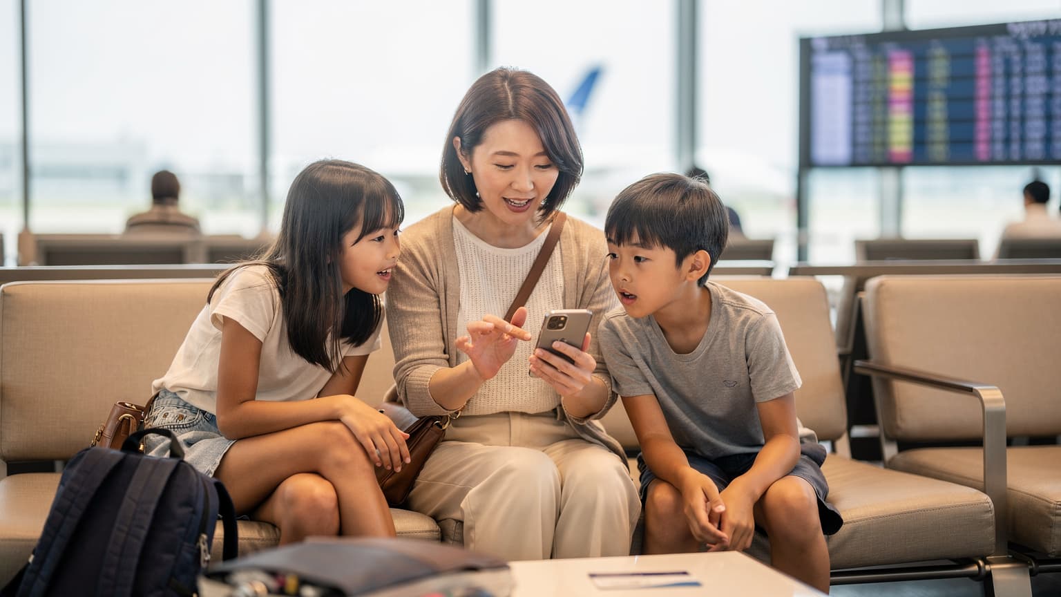 羽田空港の国際線ターミナルで、搭乗ゲートの電光掲示板を背景に、母親が子供たちと一緒にスマートフォンでeSIMの設定を確認している様子。