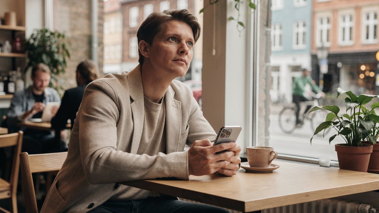 En person sidder på en travl cafe i København, kigger op fra sin iPhone 18, som viser et eSIM-installationsskærmbillede, med en dampende kaffekop ved siden af.