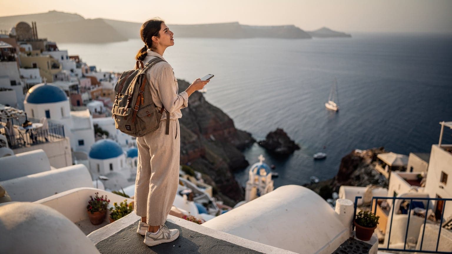 En kvinna i 20-årsåldern med ryggsäck, iklädd lätta kläder, står på en vitkalkad terrass på Santorini och tittar ut över det Egeiska havet med en smarttelefon i handen. Hon håller telefonen upp och ner, som om hon just har kollat något viktigt. Fångad under 'gyllene timmen' i Oia.