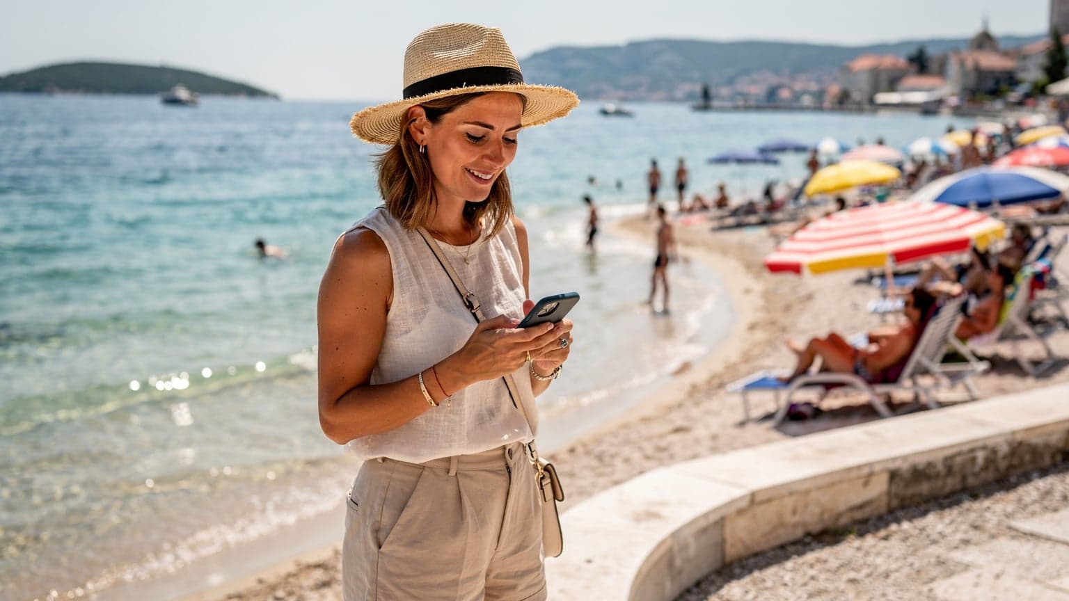 Une voyageuse souriante, téléphone en main, consulte une carte numérique sur une plage animée de la Dalmatie, avec des parasols colorés et la mer Adriatique en arrière-plan sous un soleil estival.