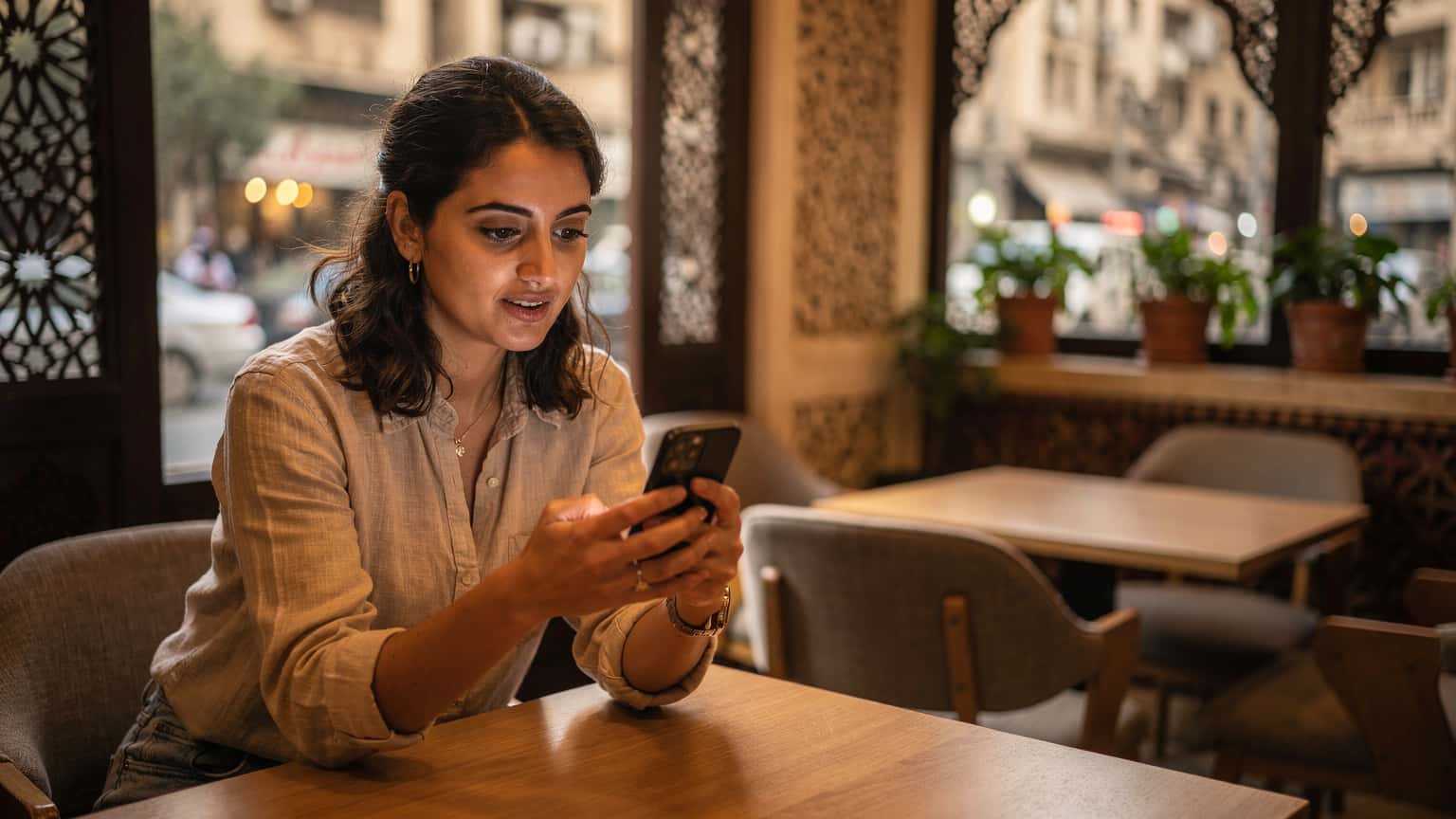 Une femme nomade numérique consultante, assise dans un café moderne du Caire avec une vue sur la ville, utilise son smartphone équipé d'une eSIM pour passer un appel vidéo, son ordinateur portable ouvert à côté d'une tasse de thé à la menthe.